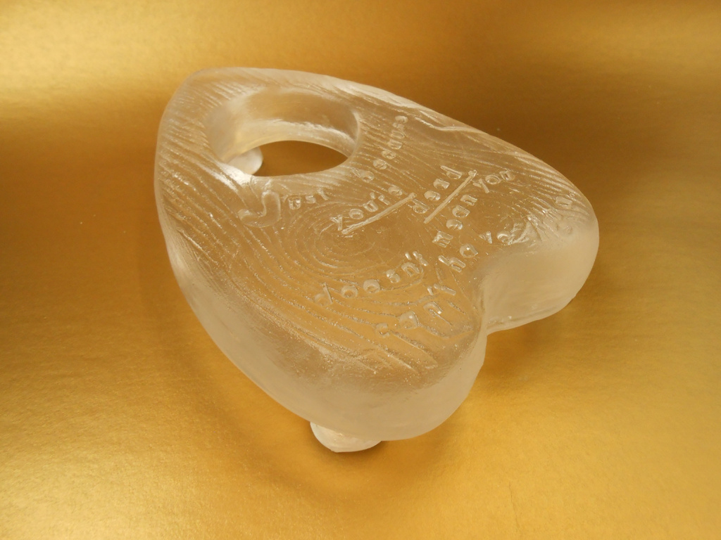 A heart-shaped planchette in textured translucent glass. The face of the planchette reads, "Just because you're dead doesn't mean you can't have fun."